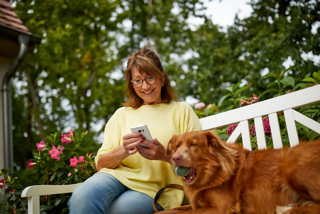 Frau sitzt auf Gartenbank und schaut auf ihr Handy. Neben ihr sitzt ein Hund
