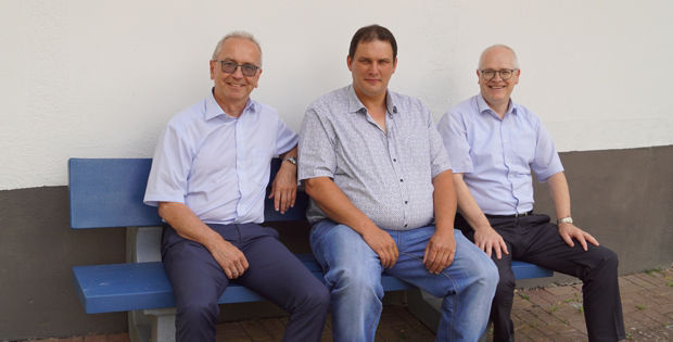 Drei Männer, alle mit hellblauen Hemden, sitzen auf einer blauen Bank vor einem weißen Gebäude.