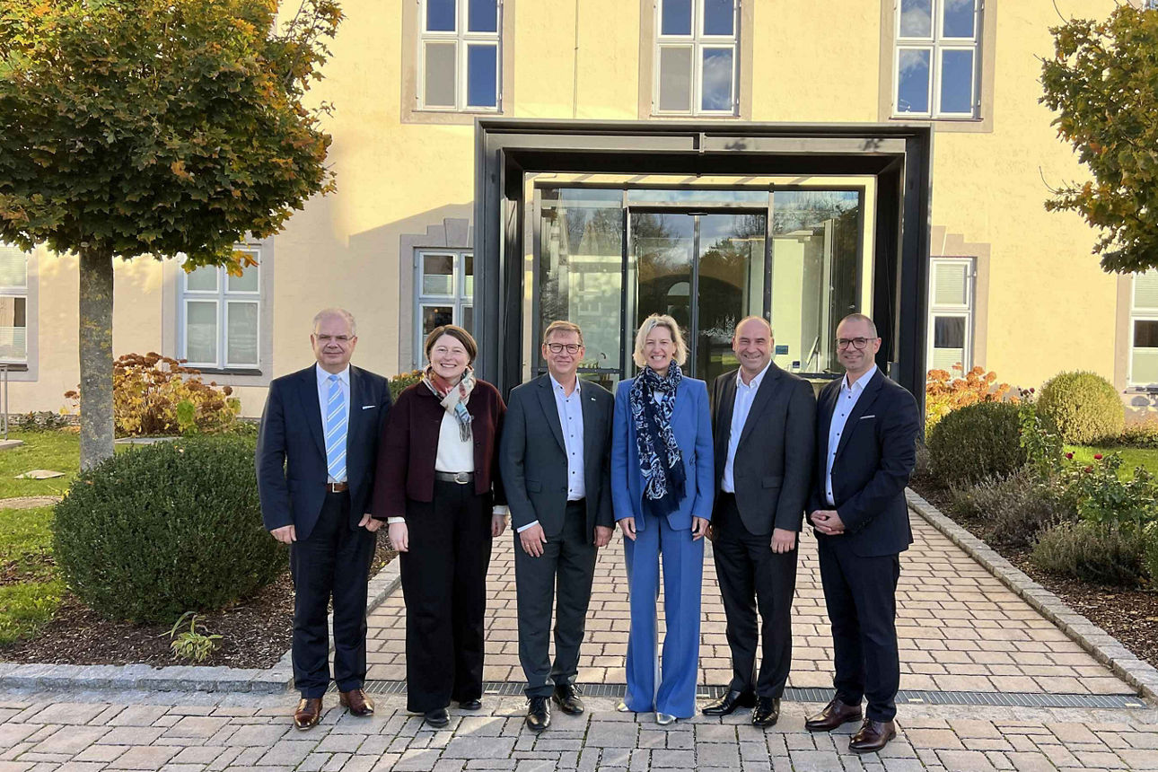 Eine Gruppe von sechs Personen steht lächelnd vor einem Gebäude und posiert für ein Foto. Alle sind formell gekleidet, offenb