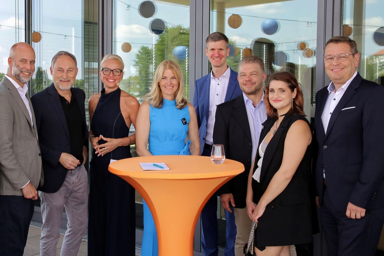 neun Personen, elegant gekleidet, stehen nebeneinander vor einer Glasfront und lächeln in die Kamera 
