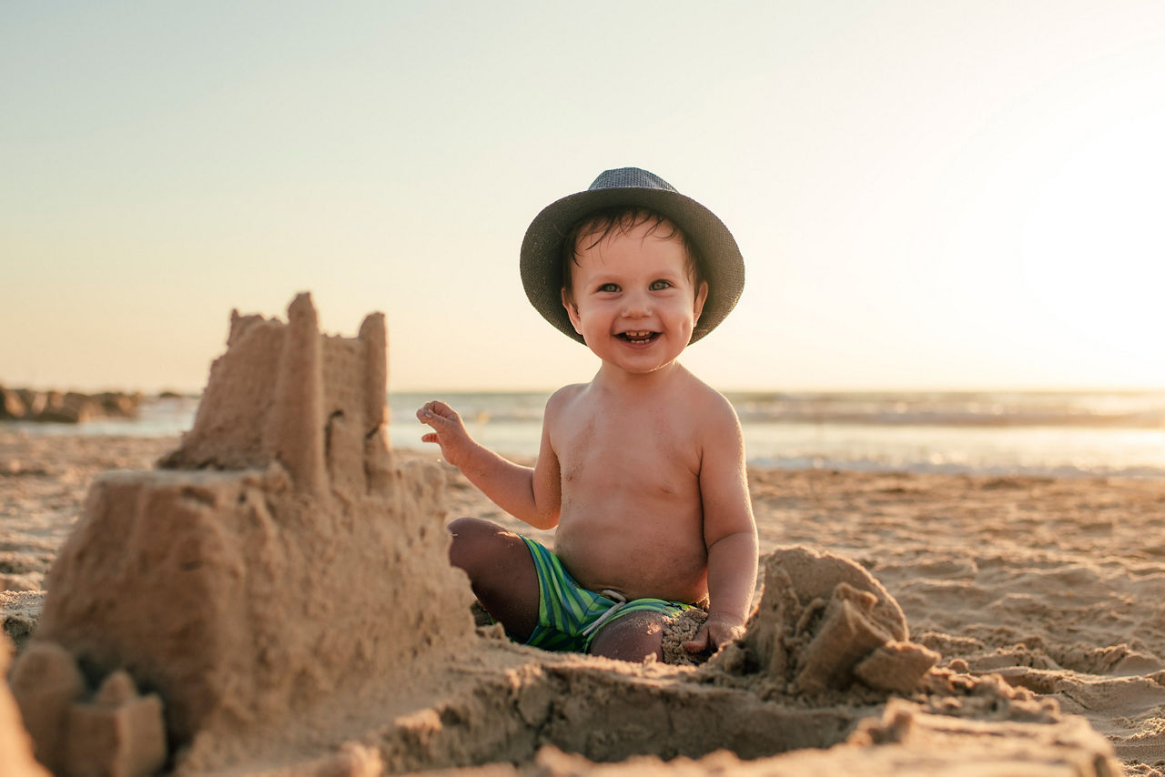 Kleinkind mit Hut sitzt am Strand im Sand