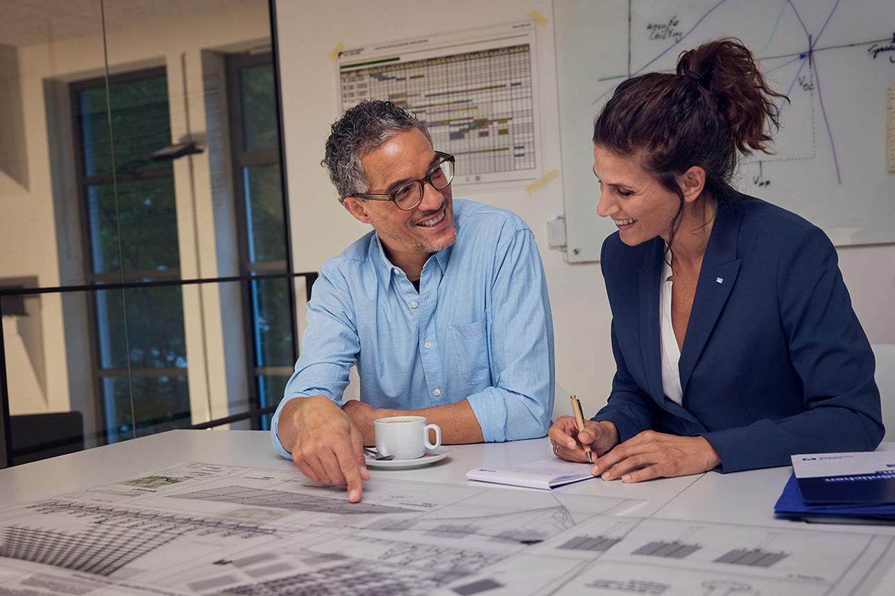 Eine Frau und ein Mann sitzen an einem Besprechungstisch und schauen sich Pläne an