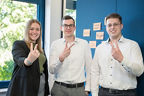 Eine Auszubildende und zwei Auszubildende stehen in einem modernen Büro und zeigen ein Volksbank-V in die Kamera