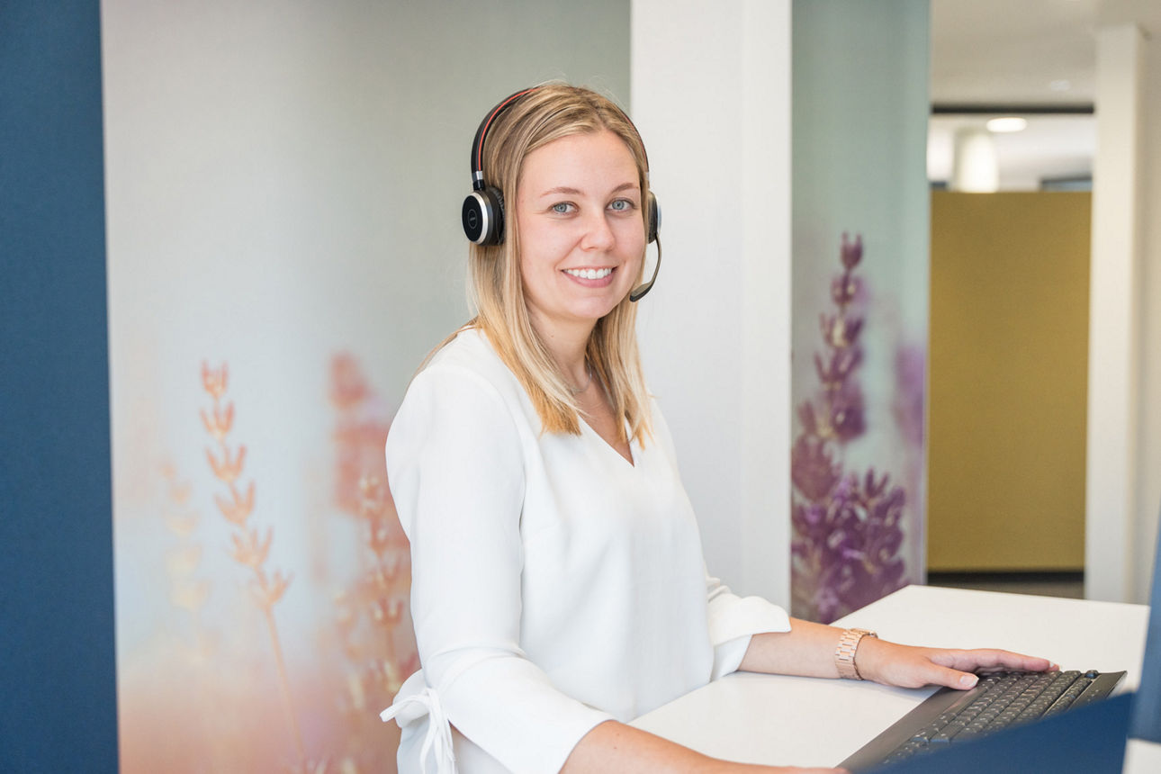 Eine Kollegin der VBBS steht mit Headset an einem Schreibtisch und lächelt in die Kamera