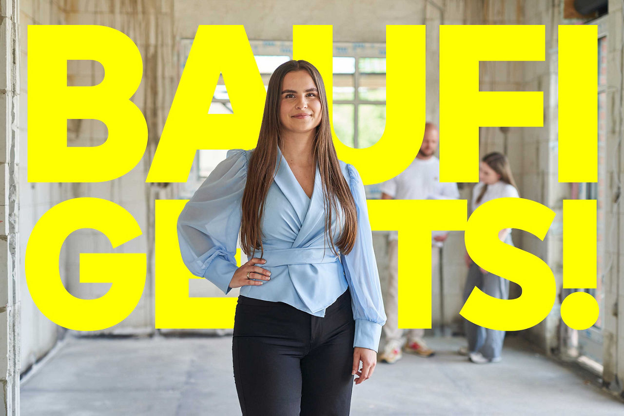 Eine junge Frau in einer hellblauen Bluse steht in einer Baustelle vor dem gelben Schriftzug "BAUFI GEHT'S!