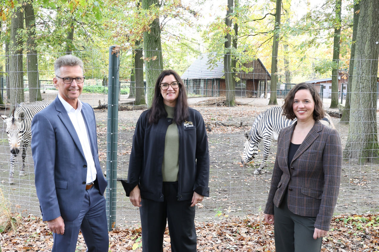 Drei Personen stehen vor einem Zebra-Gehege im Zoo und lächeln in die Kamera.