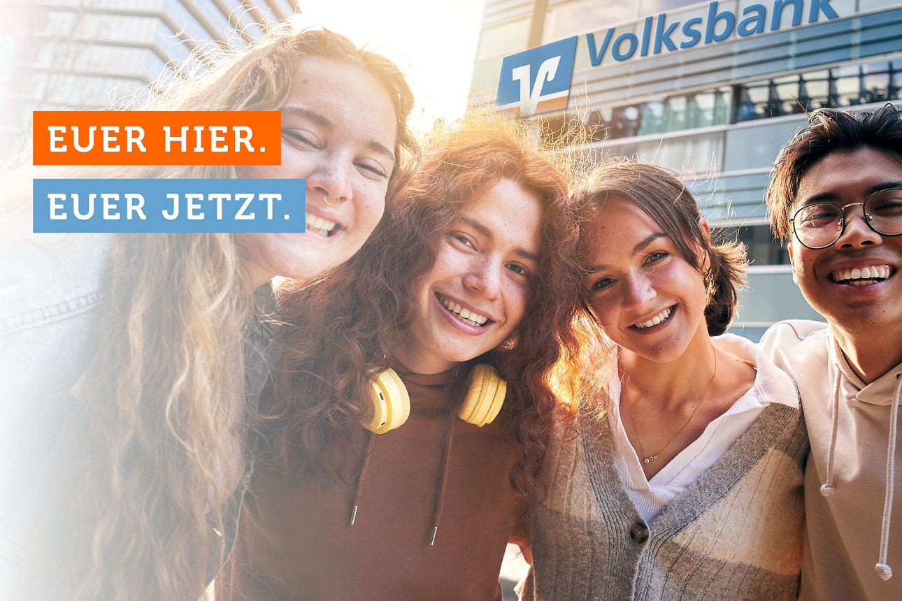 Vier junge Personen stehen vor einer Volksbank und machen ein Selfie auf dem sie lächeln.