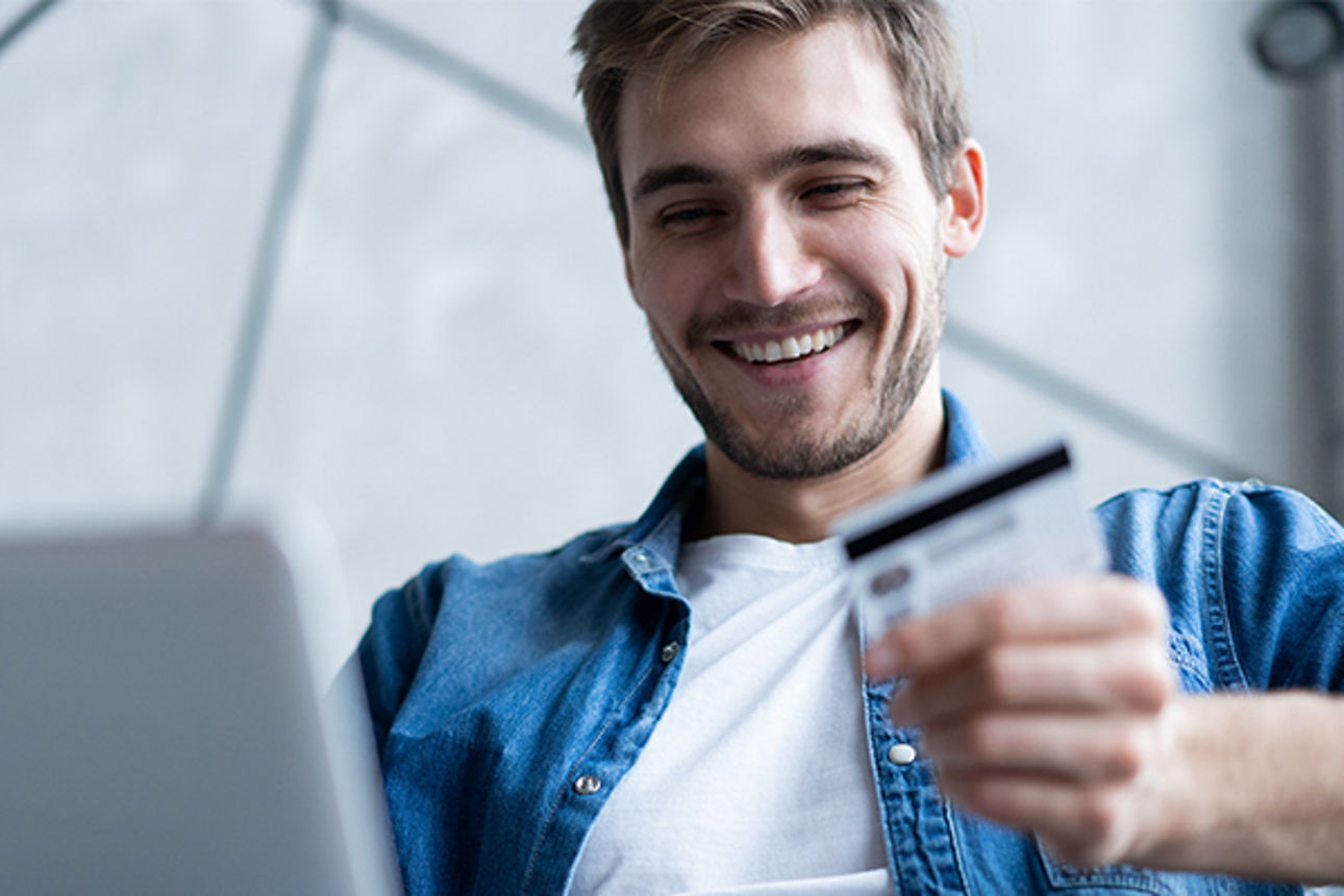 Junger Mann shoppt per Laptop im Internet, wobei er in der linken Hand eine girocard oder Kreditkarte hält 