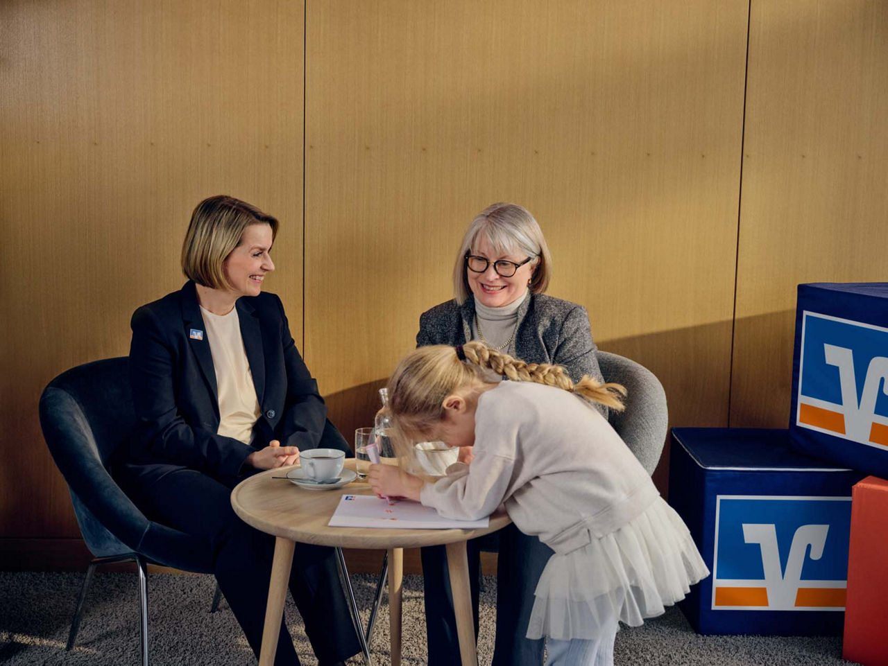 Eine Beraterin und eine ältere Frau sitzen an einem runden Tisch, daneben malt ein kleines Mädchen