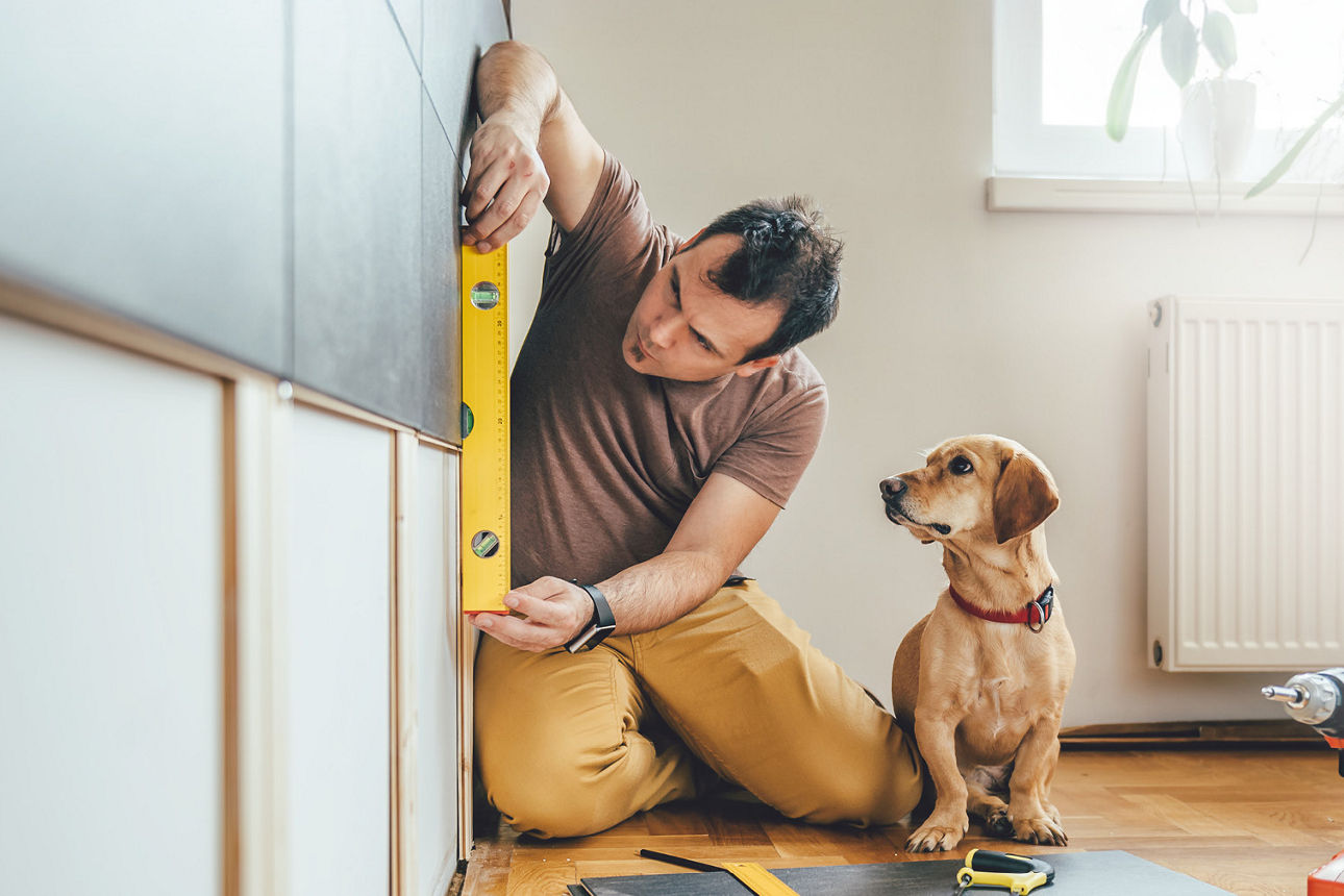 Mann knieend auf dem Boden mit Wasserwaage in der Hand und Hund sitzt daneben