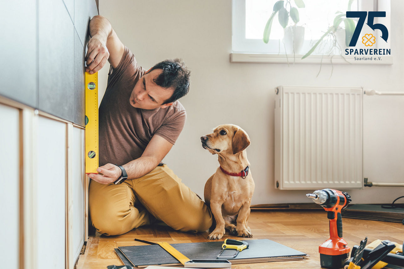 Mann knieend auf dem Boden mit Wasserwaage in der Hand und Hund sitzt daneben