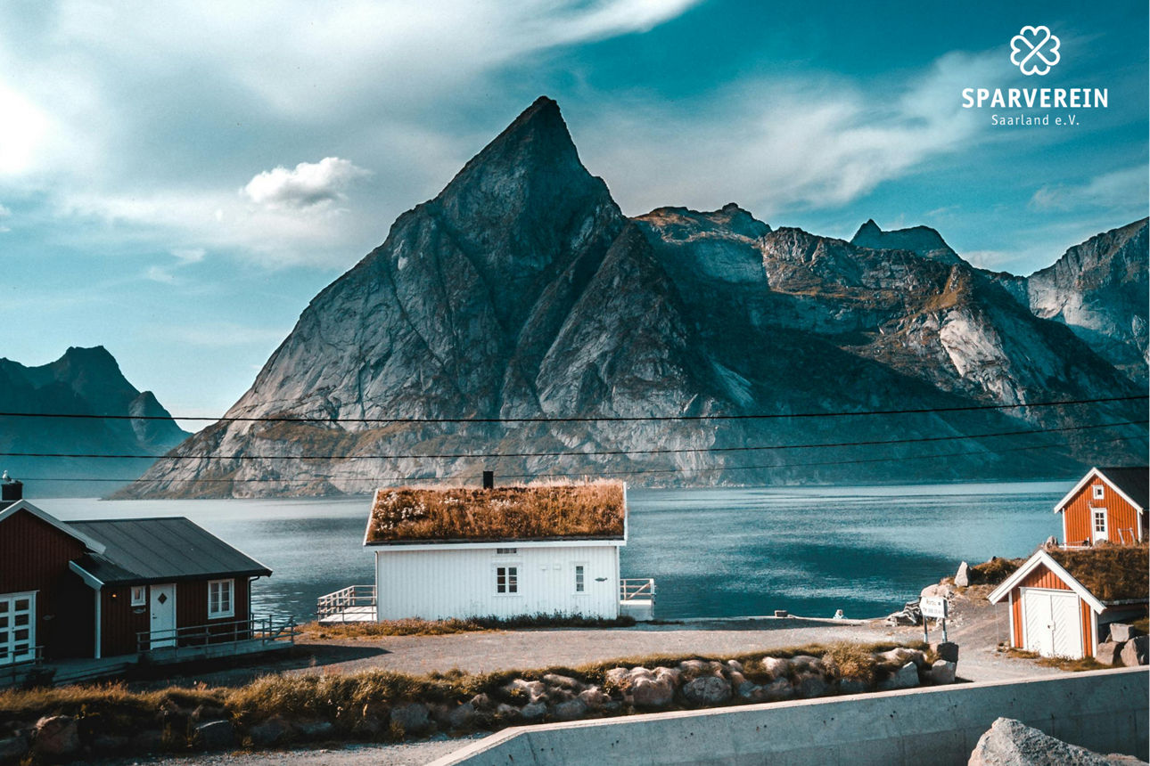 Landschaft mit Häusern vor einem Gebirge und einem See