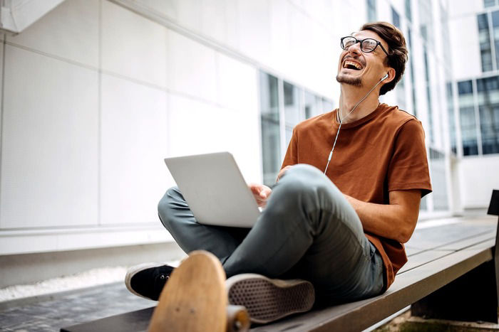 Ein junger Mann sitzt lachen auf einer Bank mit einem Laptop in einem Innenhof
