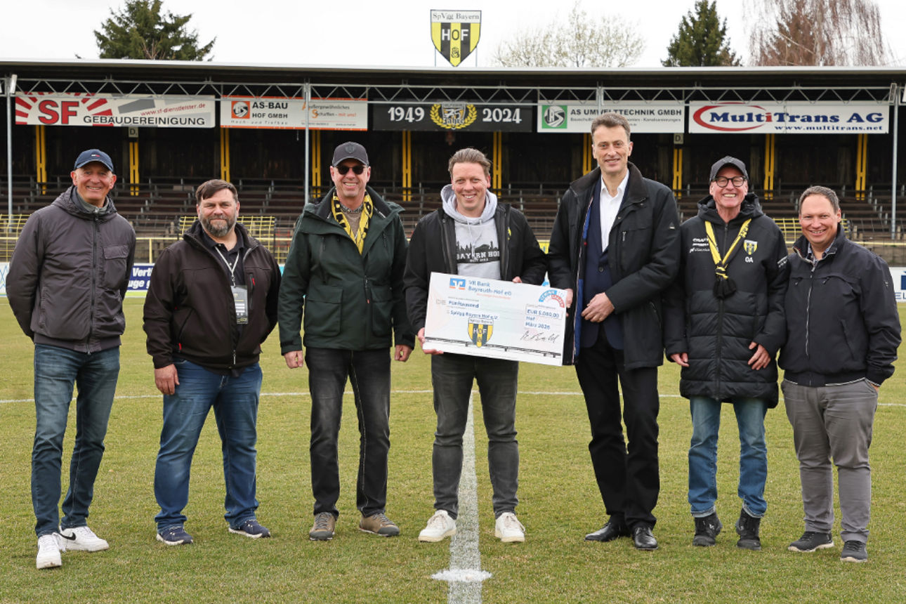 Gruppenfoto auf einem Fußballfeld, bei dem mehrere Personen gemeinsam einen großen symbolischen Spendenscheck präsentieren.