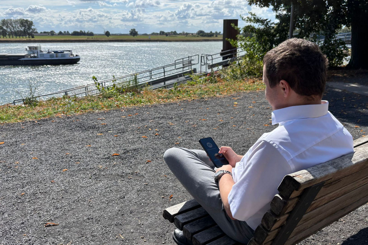 Junger Mann sitzt an einem Fluss und schaut auf sein Smartphone