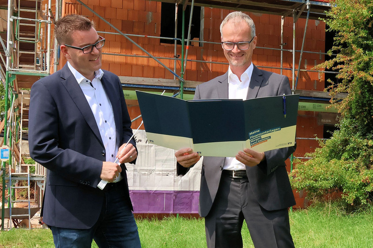 Kundenberater Ulrich Gagstetter und Jürgen Hertlein stehen vor Rohbau und betrachten einen Plan