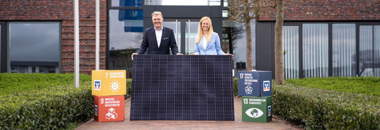 Eine Frau und ein Mann halten einen Photovoltaikpaneel in  der Hand und stehen vor einem großen Gebäude