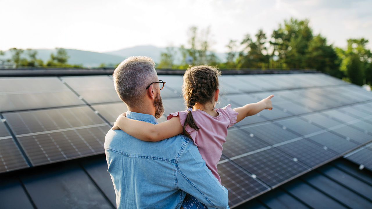 Mann mit Kind auf dem Arm schaut auf ein Hausdach mit Photovoltaikanlage