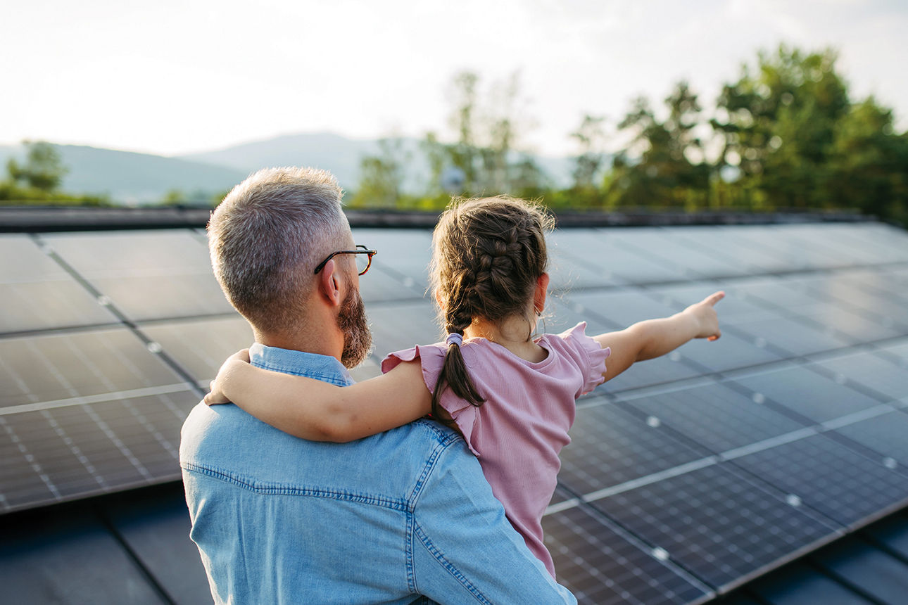 Mann mit Kind auf dem Arm schaut auf ein Hausdach mit Photovoltaikanlage