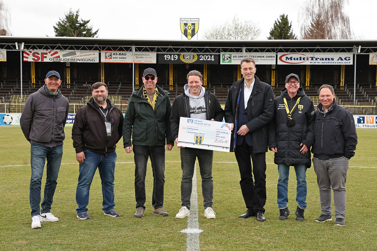 Gruppenfoto auf einem Fußballfeld, bei dem mehrere Personen gemeinsam einen großen symbolischen Spendenscheck präsentieren.
