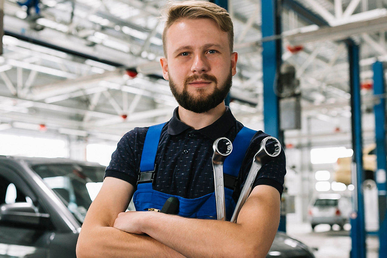 Ein Kfz-Mechaniker mit Bart steht mit verschränkten Armen und Schraubenschlüsseln in einer Werkstatt.