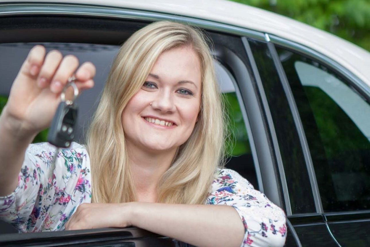 Eine junge Frau hält lächelnd einen Autoschlüssel aus der Fahrerseite des Autos.
