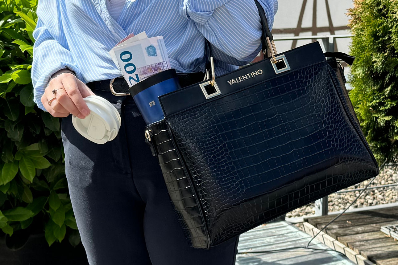 Jemand hält eine Handtasche darin ist ein To-Go Becher zu sehen, in welchem Geldscheine stecken.