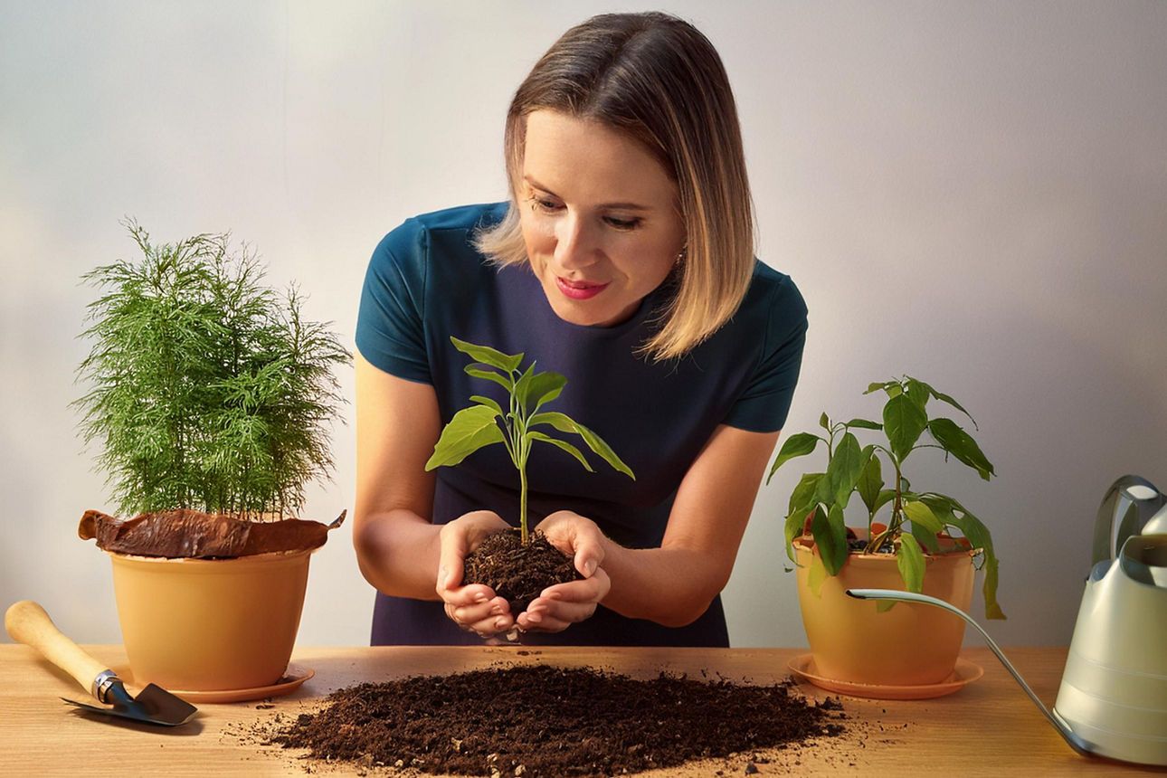 Eine Frau hält eine Pflanze in den Händen, darunter ist Erde auf einem Tisch, daneben stehen Töpfe mit größeren Pflanzen.