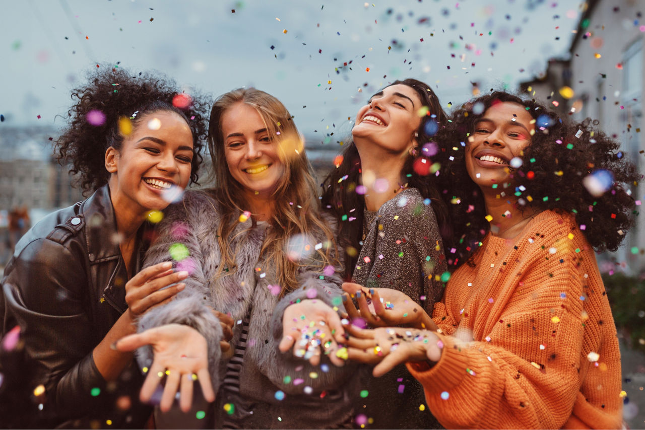 Vier junge Frauen lachen und feiern draußen mit buntem Konfetti in der Luft, fröhliche und ausgelassene Stimmung.