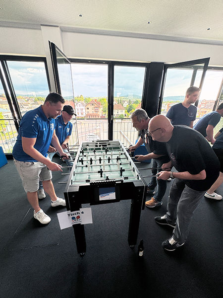 Auf dem Bild ist ein Tischkicker zu sehen. Links spielen zwei Spieler in blauem und rechts in schwarzem Shirt.