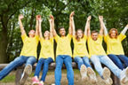 Sechs Erwachsene in gelben Shirts jubeln. Sie streck ihre Hände in die Luft und sitzen nebeneinander auf einem Baumstamm.
