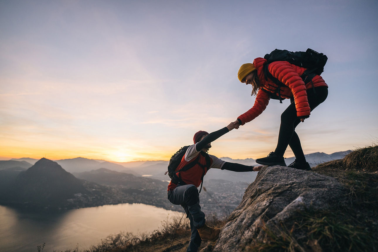 Eine Frau reicht ihrem Partner beim Bergsteigen die Hand - man blickt auf Berge, Sonnenaufgang und See