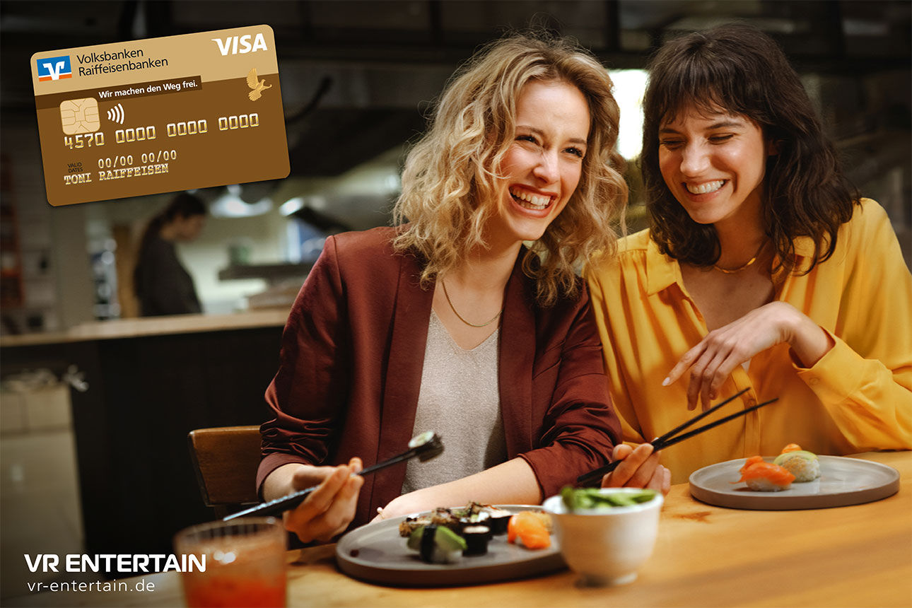 Zwei Frauen sitzen an einem Tisch mit Sushi, eine trägt ein rotes Jackett, die andere ein gelbes Oberteil. Oben links ist ein