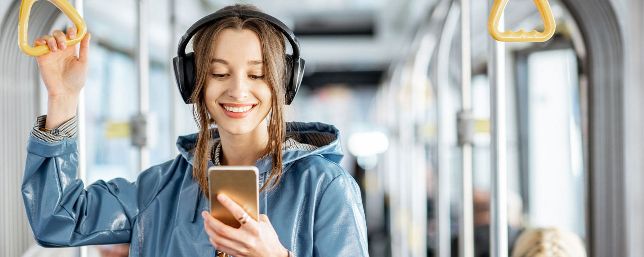 Junge Frau mit Kopfhörer steht im Bus und schaut auf ihr Handy