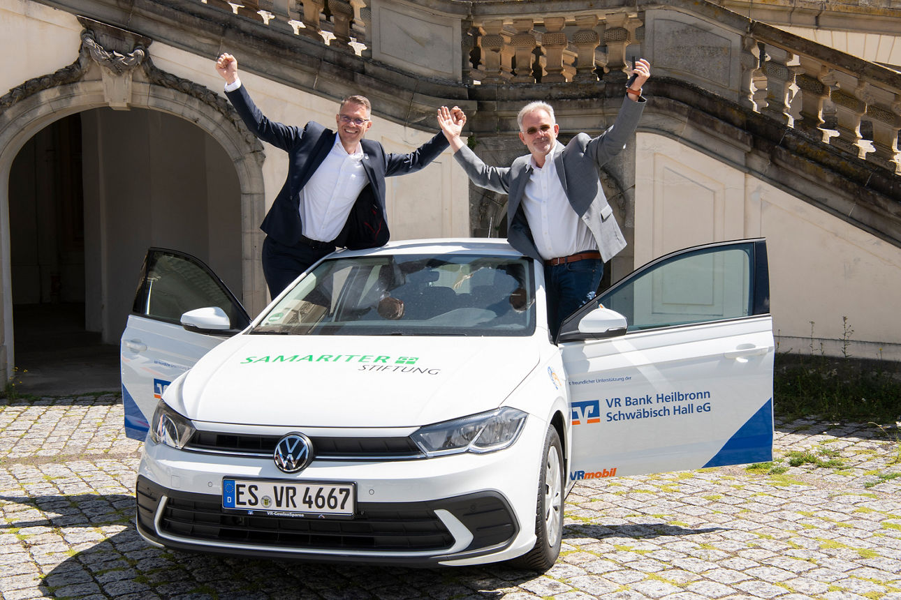Zwei Männer stehen mit erhobenen Armen freudig aus den offenen Vordertüren eines weißen VW-Pkw. Auf der Motorhaube steht „Samariter Stiftung“, an der Seite „VR Bank Heilbronn Schwäbisch Hall eG“. Das Auto steht auf einem gepflasterten Platz.