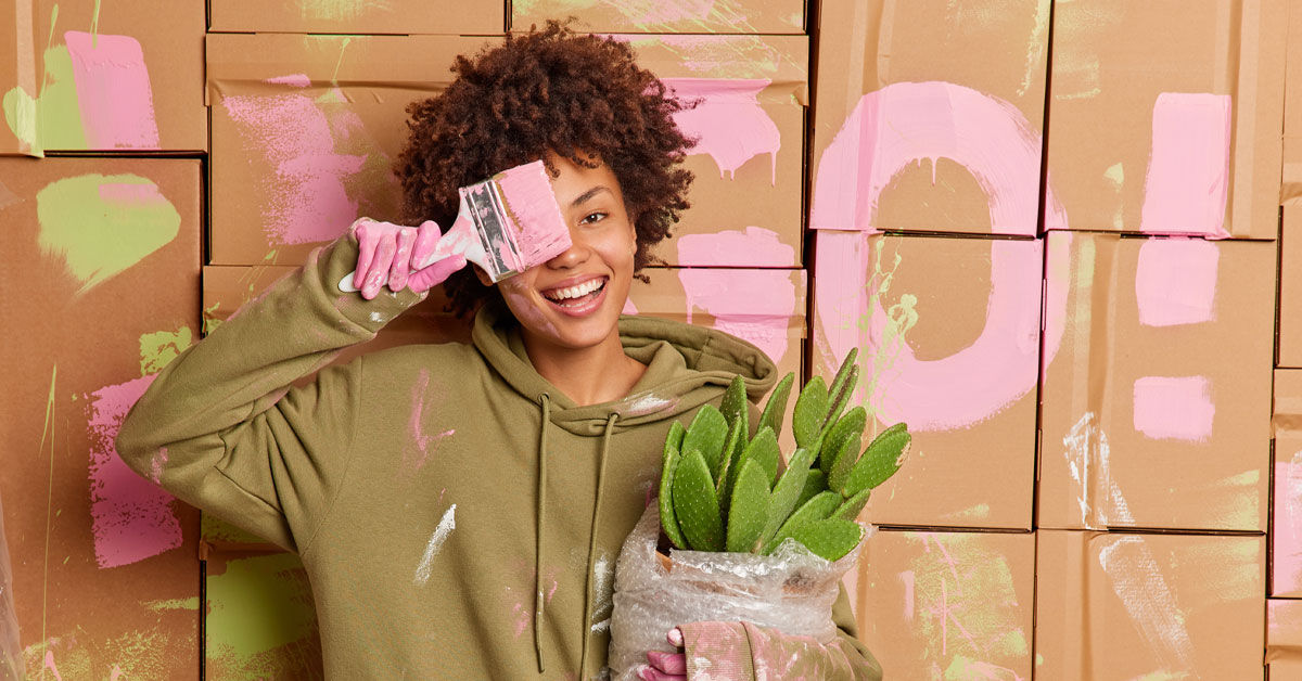 Eine Frau, die eine Blume in der Hand hält und sich einen Pinsel mit pinker Farbe vor das rechte Auge hält