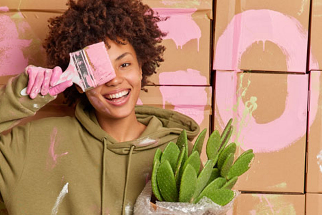 Eine Frau, die eine Blume in der Hand hält und sich einen Pinsel mit pinker Farbe vor das rechte Auge hält