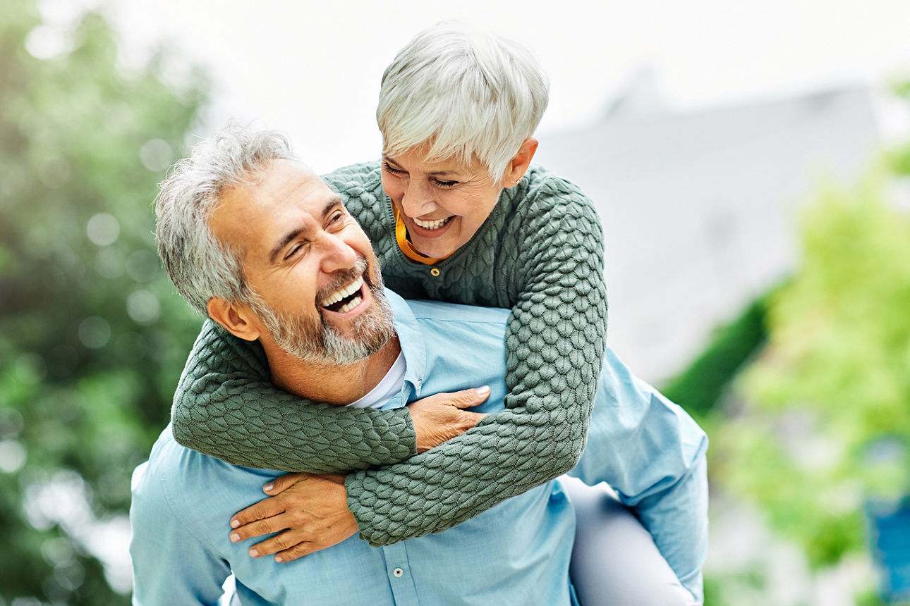 Ältere Person trägt eine weitere Person mit grauen Haaren auf dem Rücken, beide tragen helle Oberteile, im Hintergrund unscharfe Garten- oder Parkanlage.