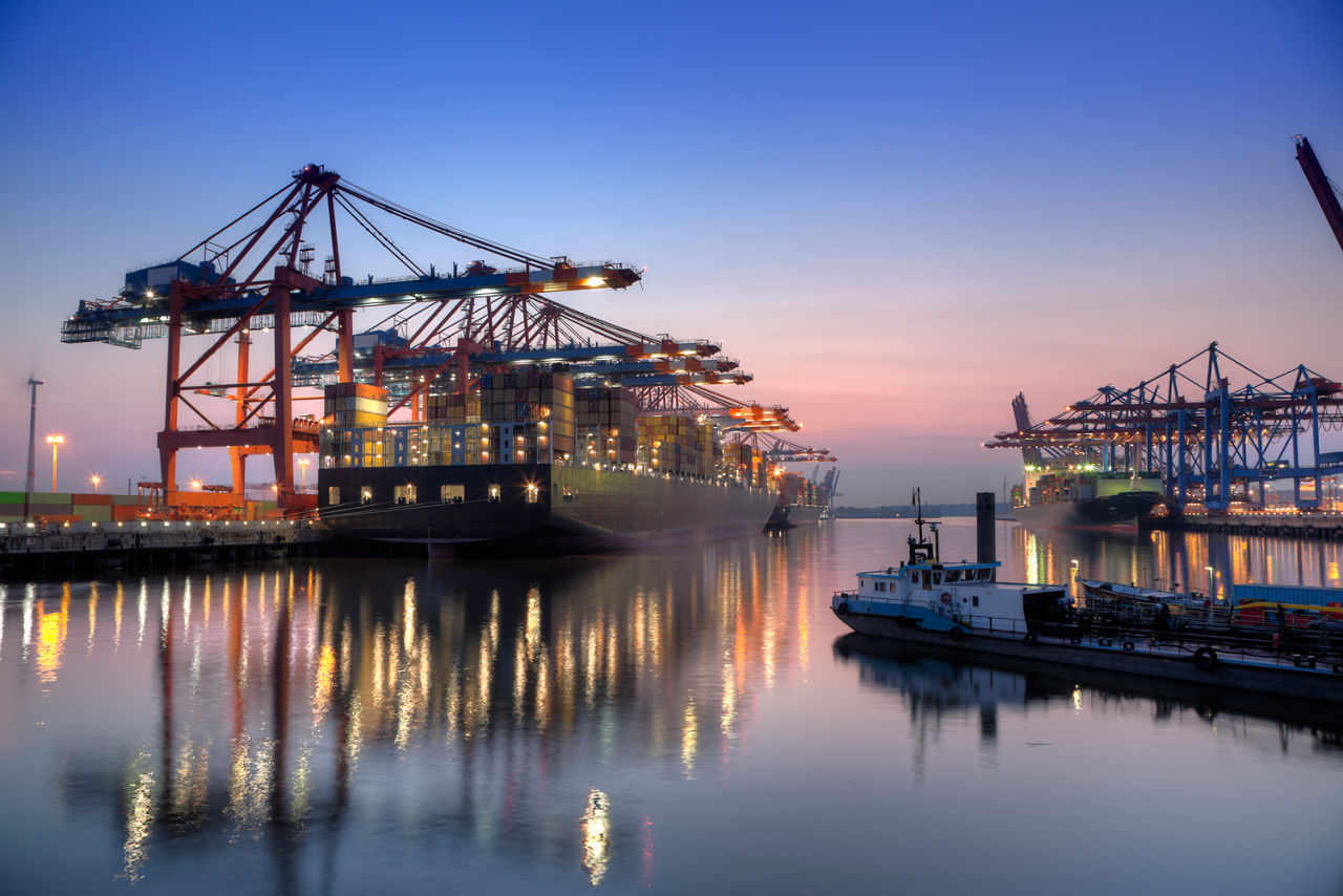 Containerterminal mit mehreren Kränen und großen Containerschiffen im Hafen bei Sonnenuntergang, reflektiert im Wasser.