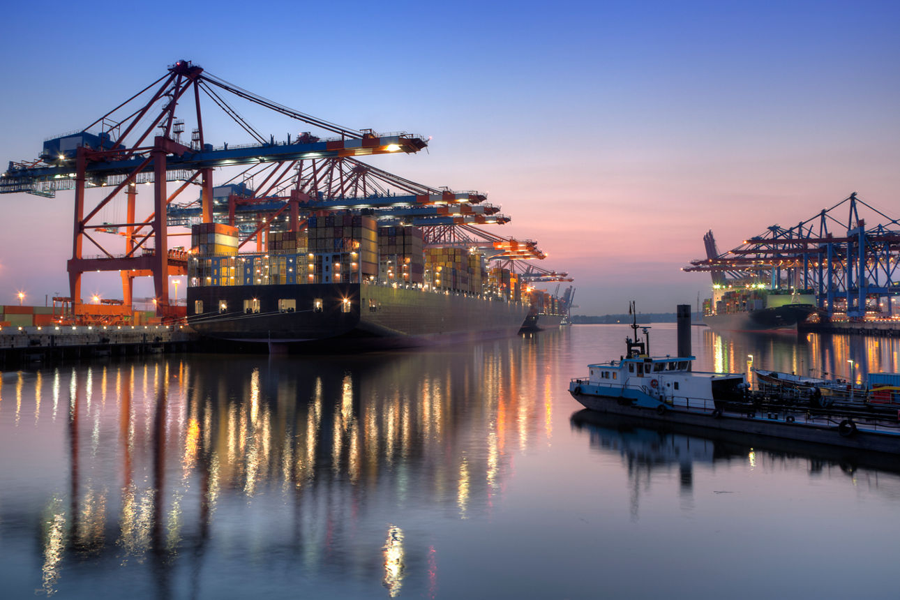 Containerterminal mit mehreren Kränen und großen Containerschiffen im Hafen bei Sonnenuntergang, reflektiert im Wasser.