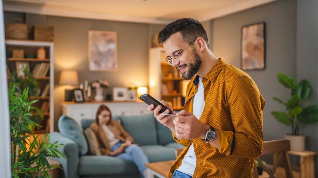 Junger Mann mit Brille hält sowohl eine Bankkarte als auch sein Handy in seiner Wohnung in der Hand
