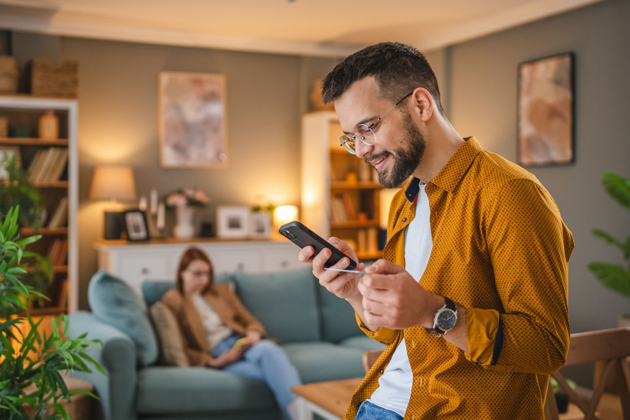 Junger Mann mit Brille hält sowohl eine Bankkarte als auch sein Handy in seiner Wohnung in der Hand