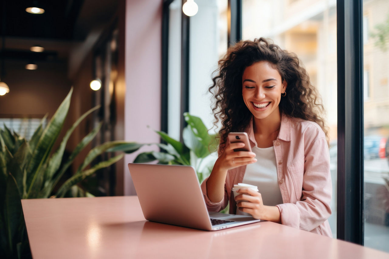 Junge Frau mit einem Becher Kaffee sitzend an Ihrem Laptop mit Handy in der Hand in einem Café