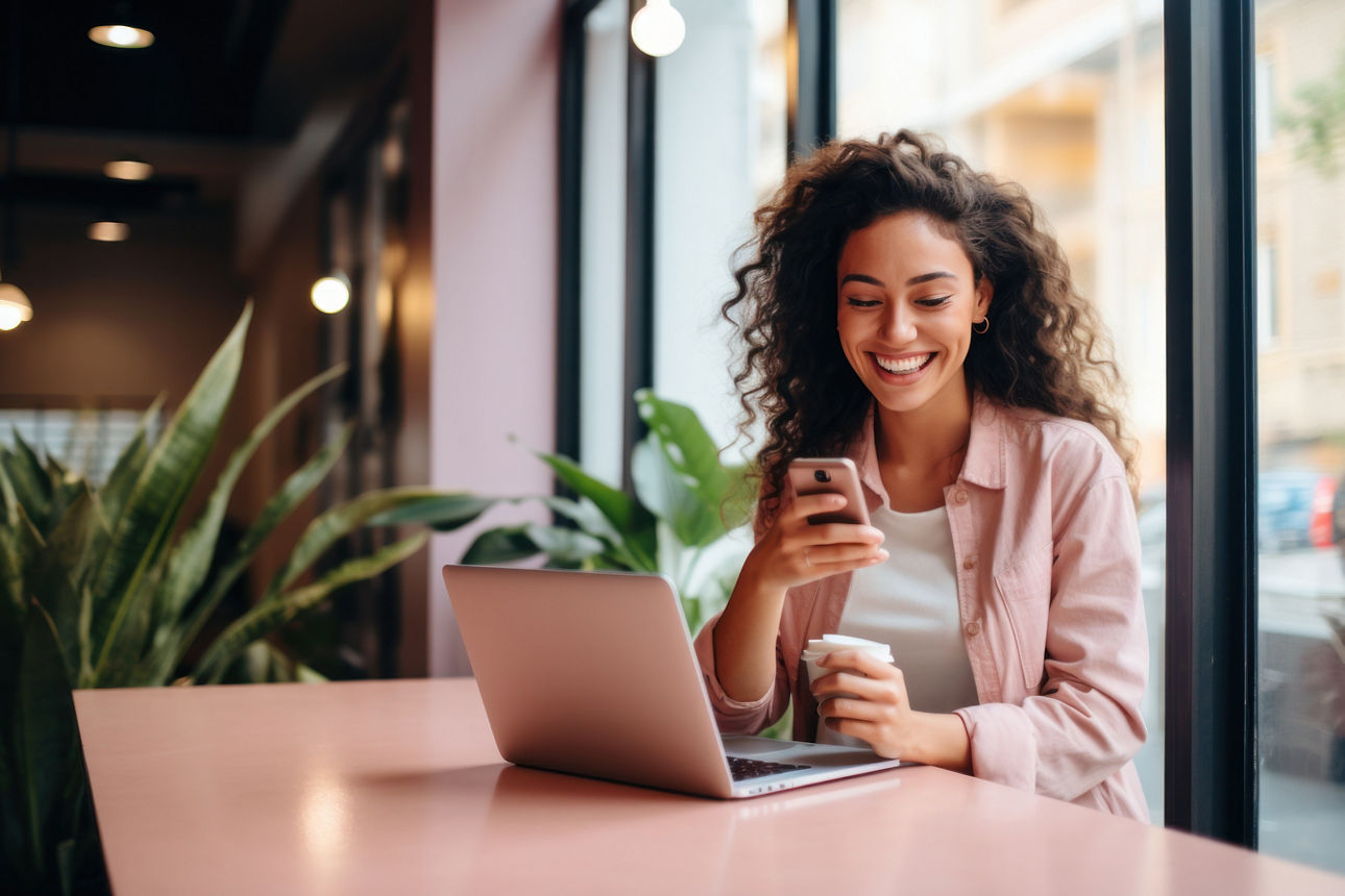 Junge Frau mit einem Becher Kaffee sitzend an Ihrem Laptop mit Handy in der Hand in einem Café