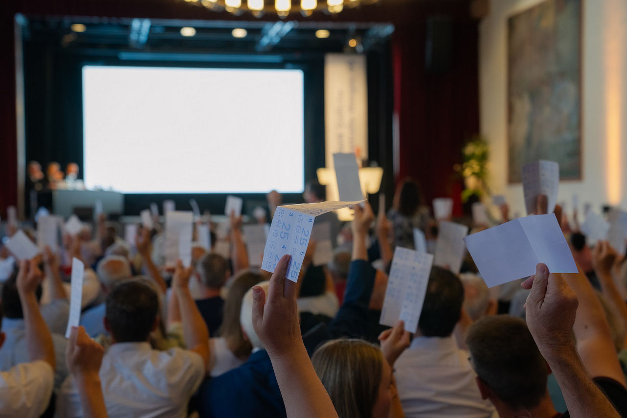 Menschen in einem Saal heben Stimmkarten bei einer Versammlung mit Bühne und Leinwand im Hintergrund.