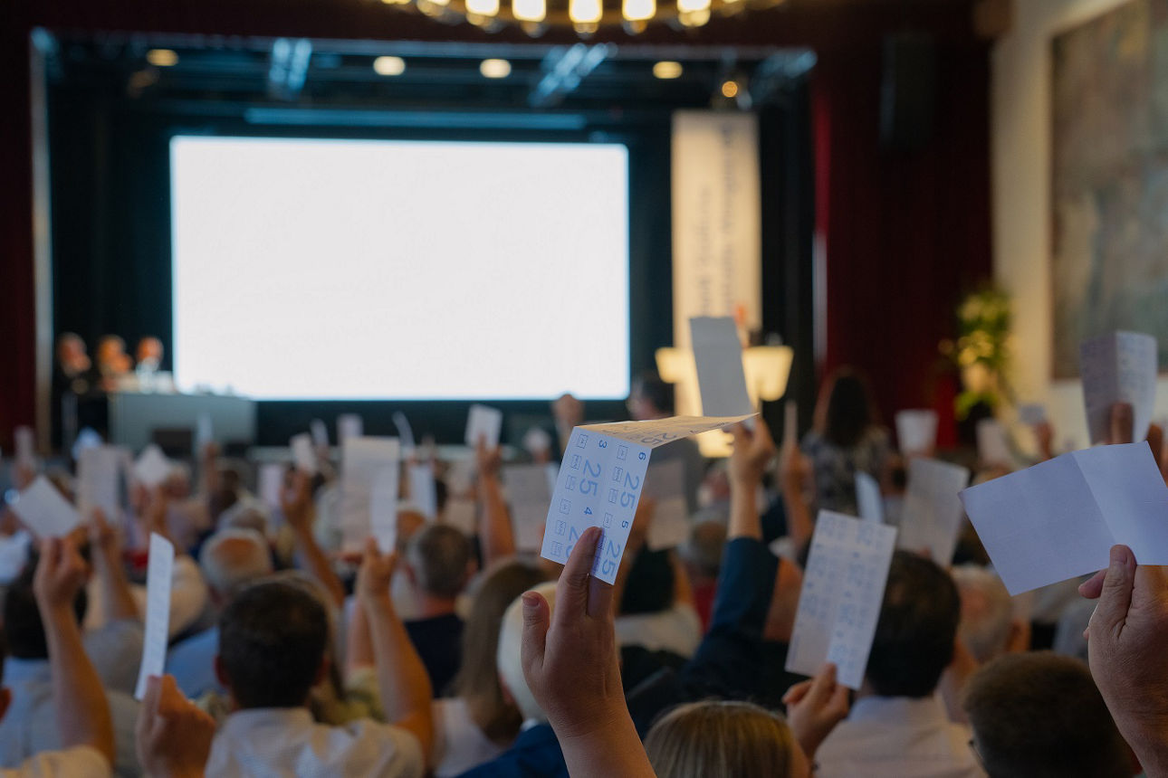 Menschen in einem Saal heben Stimmkarten bei einer Versammlung mit Bühne und Leinwand im Hintergrund.