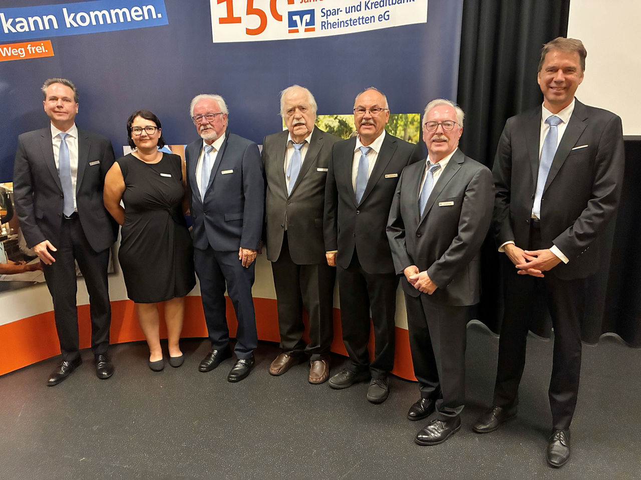 Eine Gruppe von Menschen, die vor einer Firmenkulisse der Spar- und Kreditbank Rheinstetten eG in einer Reihe stehen.