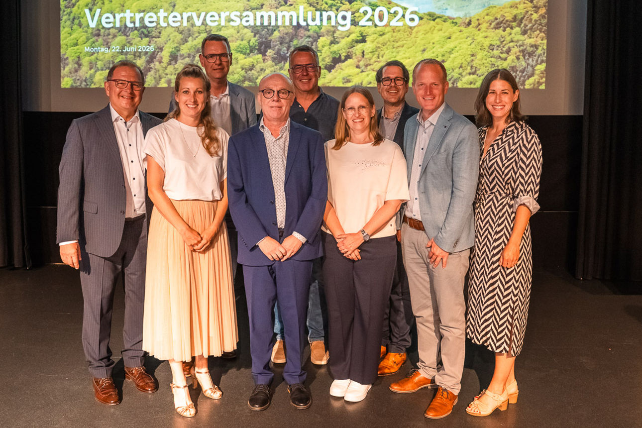 Gruppe von Personen steht auf Bühne vor Leinwand mit Text ‚Herzlich willkommen Vertreterversammlung 2026
