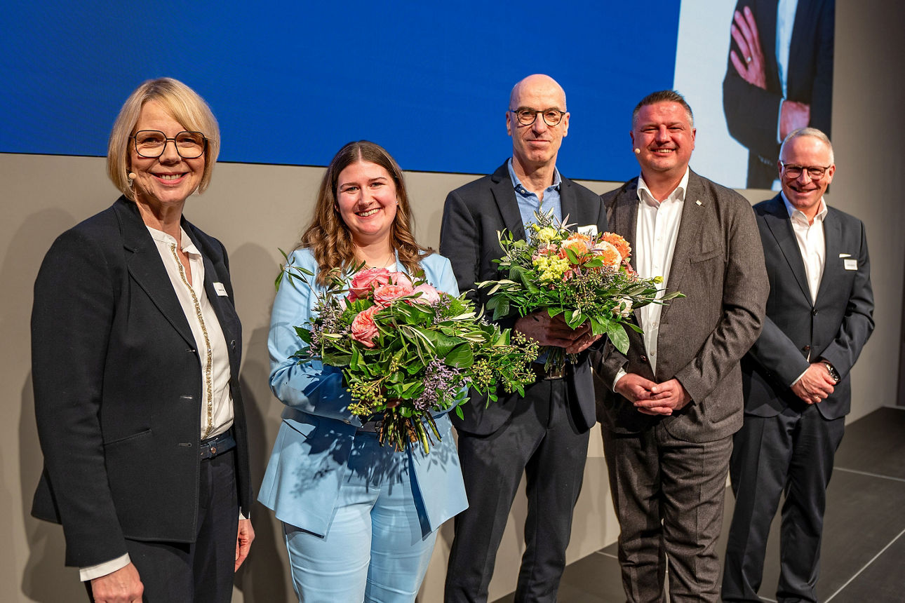 Ehrung auf der Bühne: Fünf Personen stehen zusammen, zwei davon halten Blumensträuße als Zeichen der Anerkennung in den Hände