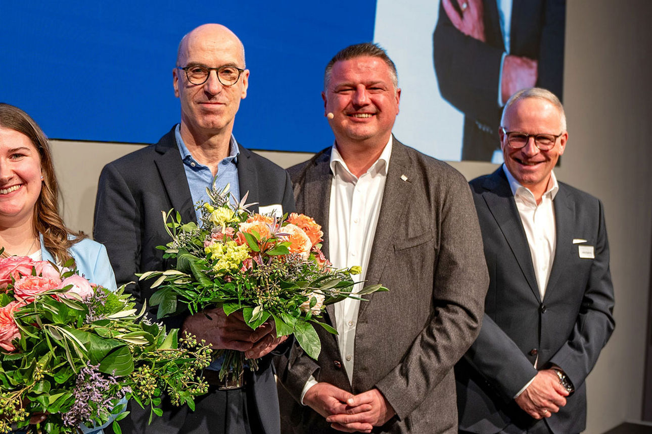 Mehrere Personen stehen bei einer Ehrung zusammen auf der Bühne und halten lächelnd bunte Blumensträuße.